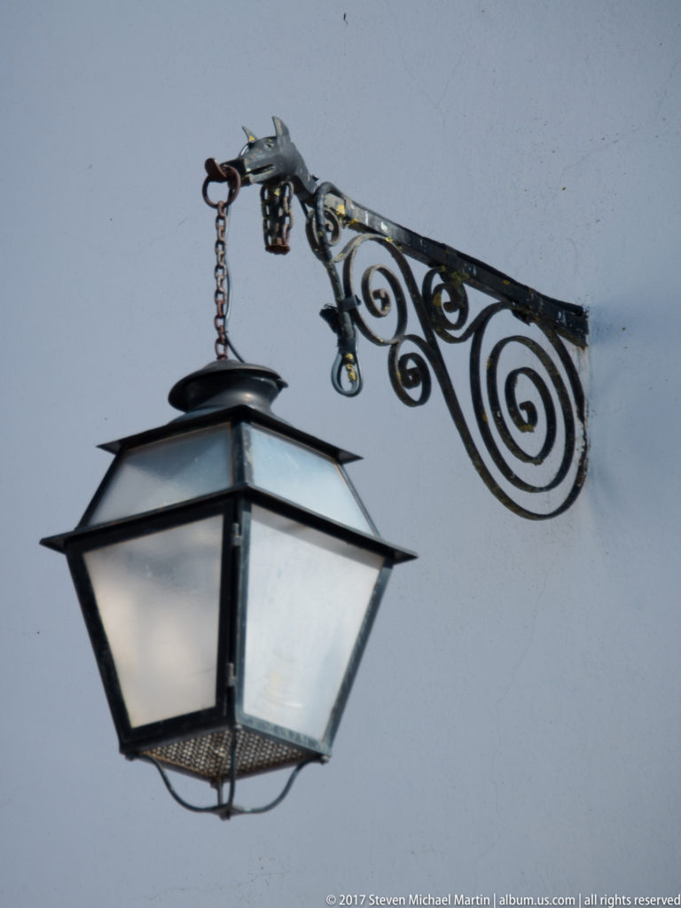 Black hanging lantern suspended by a chain from an ornate scrolling wrought-iron bracket on a white wall.