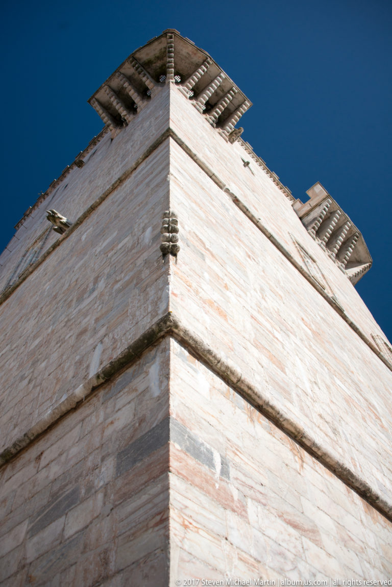 Low-angle close view of a pale stone fortified tower corner with projecting corbels against a deep blue sky.