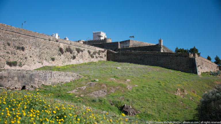 Long stone fortification walls climbing a green hillside toward white castle buildings under a clear blue sky.