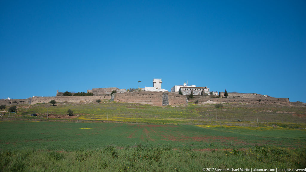 Distant fortified hilltop with castle keep and white buildings beyond broad green fields dotted with yellow flowers.