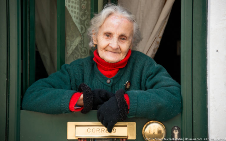 Close portrait of an elderly woman at a green door, with gloved hands beside a brass mail slot marked CORREIO.