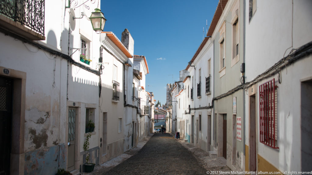 Narrow cobbled street descending between whitewashed houses with tiled roofs, balconies, lamps, and colorful window trim.