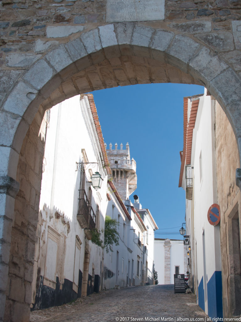 Whitewashed street and distant crenellated tower framed by a broad stone arch.