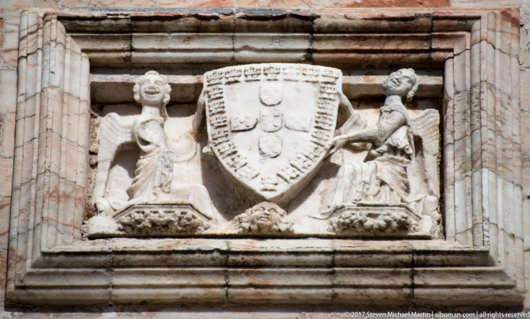 Weathered stone relief of the Portuguese royal shield flanked by two carved figures within a molded frame.