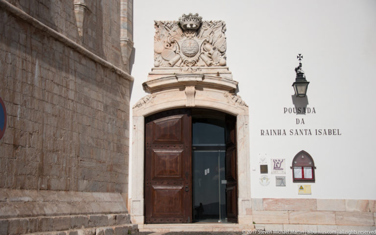 Stone-framed wooden entrance beneath a carved coat of arms and the words POUSADA DA RAINHA SANTA ISABEL.