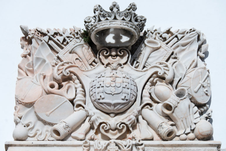 Ornate pale stone Portuguese coat of arms surrounded by crown, scrollwork, flags, armor, and military trophies.