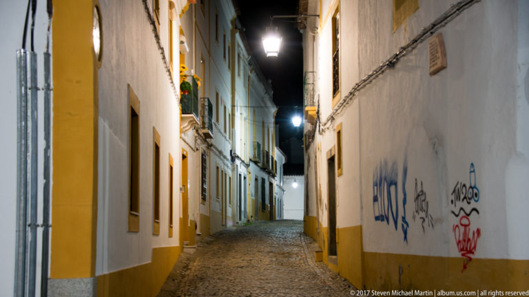 Narrow cobbled Évora lane at night between whitewashed buildings, with ochre trim, hanging street lamps, and graffiti.