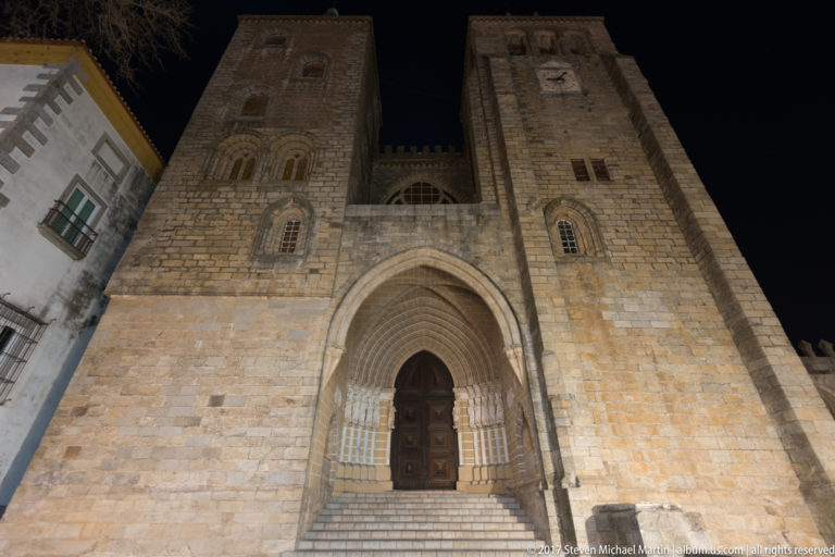 Low-angle night view of Évora Cathedral's twin-towered west façade with central Gothic portal and broad stone stair.