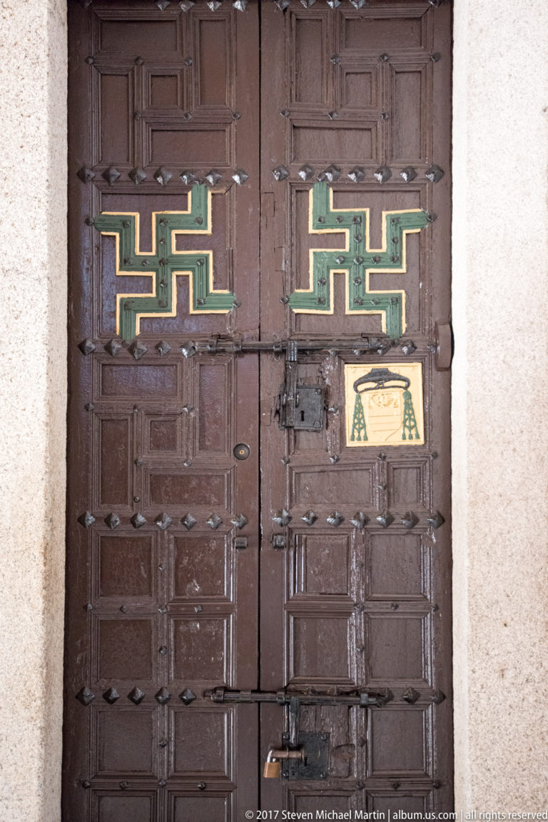 Heavy studded wooden double doors with paired green-and-gold geometric motifs and a small painted panel near the lock.