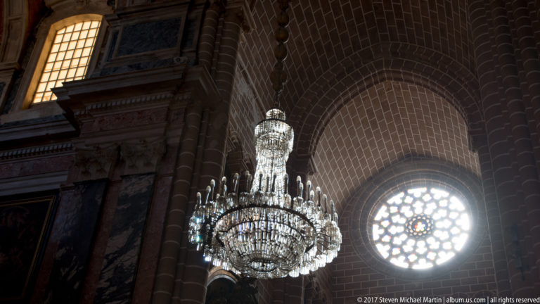 Backlit crystal chandelier beside a glowing stained-glass rose window under dark stone vaulting in Évora Cathedral.