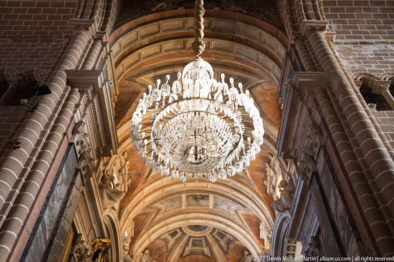 Crystal chandelier suspended beneath a richly painted vault between massive reddish stone piers in Évora Cathedral.