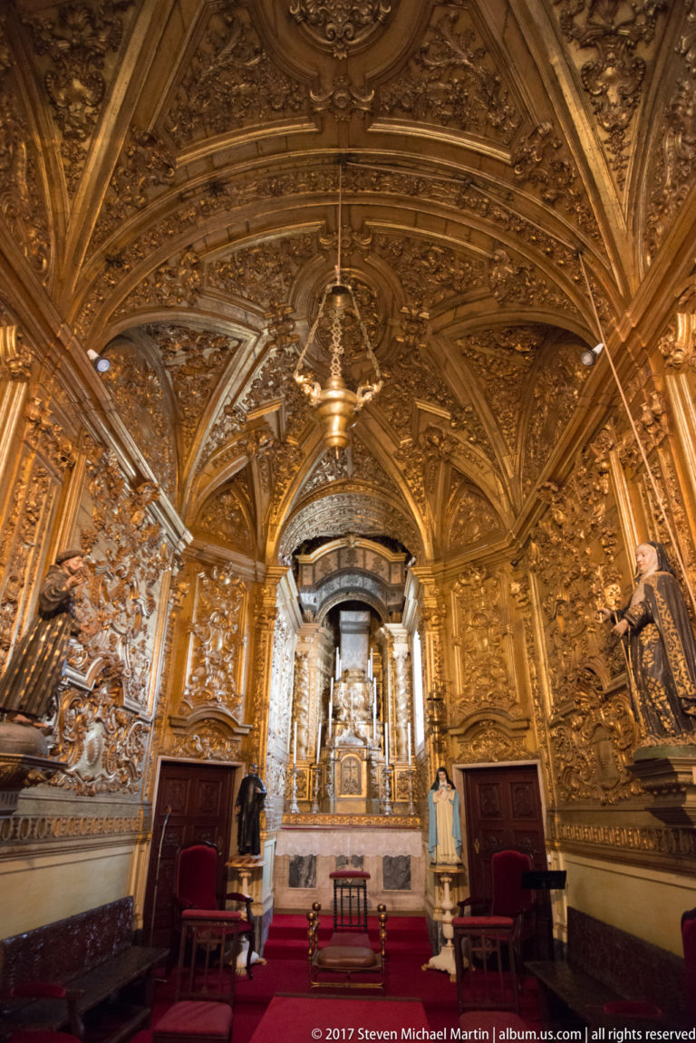 Small Baroque chapel covered in gilded carved decoration, with altar, red doors, chairs, and richly ornamented vault.