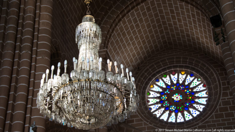 Large crystal chandelier hanging before a stained-glass rose window beneath dark stone vaults in Évora Cathedral.