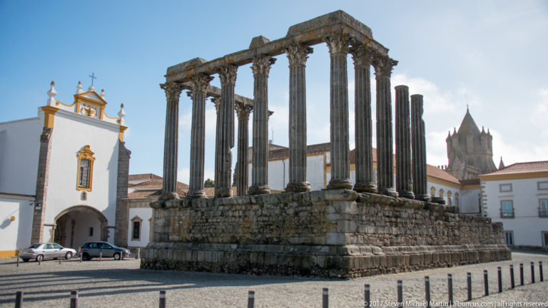 Roman Temple of Évora with tall granite Corinthian columns on a raised stone podium beside white historic buildings.