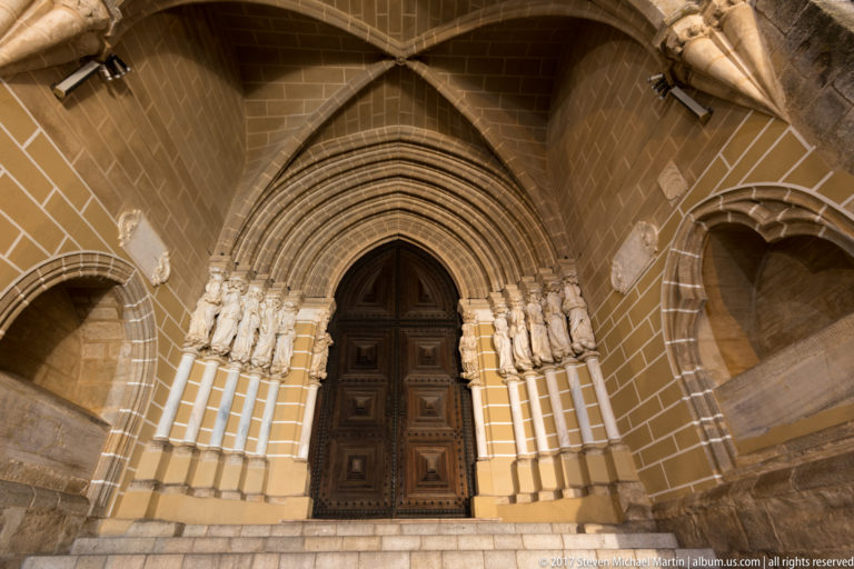 Illuminated Gothic portal of Évora Cathedral with carved apostles, pointed archivolts, wooden doors, and ribbed porch vault.