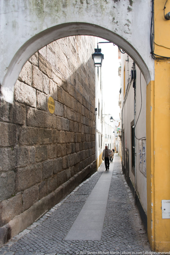 Narrow cobbled lane under an arch, bordered by a massive stone wall and whitewashed houses with pedestrians beyond.