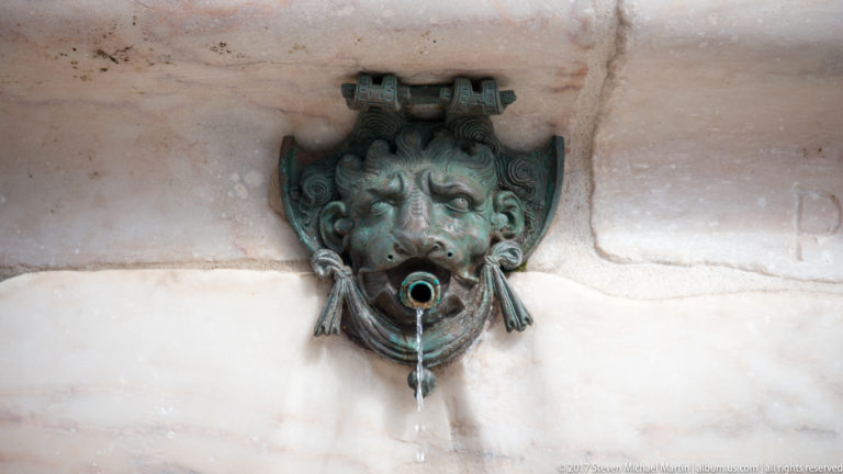 Patinated bronze face-shaped fountain spout releasing water against a pale stone wall.