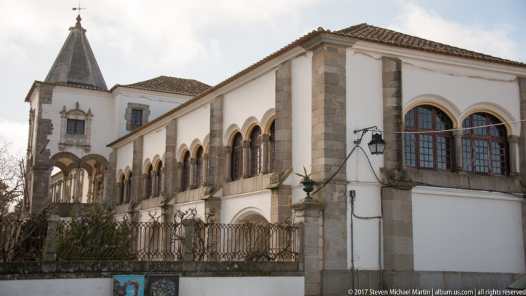 Oblique view of a long white historic façade with stone arches, glazed openings, and granite corner details.
