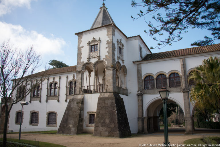 White historic complex with square tower, stone-trimmed windows, elevated arcade, and large arched passage.