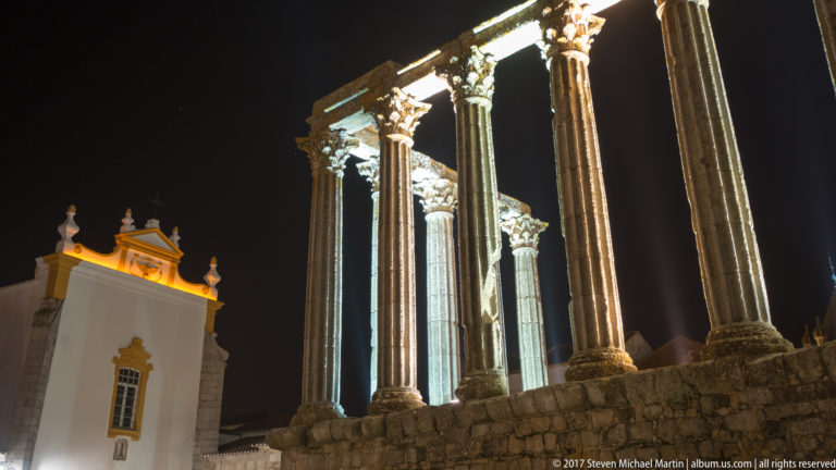 Oblique night view of the Roman Temple of Évora beside a white-and-ochre historic building.