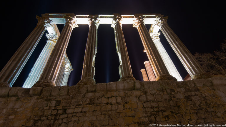 Low-angle night view of the Roman Temple of Évora with floodlit Corinthian columns above a rough stone podium.