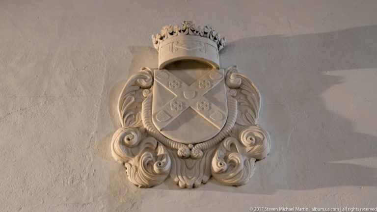 Crowned carved-stone coat of arms with diagonal cross and scrolling ornamental surround on a pale wall.