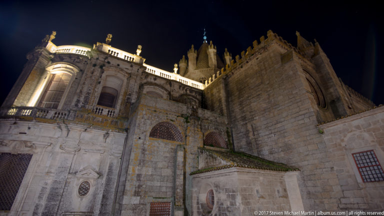 Floodlit stone exterior of Évora Cathedral at night, showing layered apse structures, buttresses, crenellations, and pinnacles.