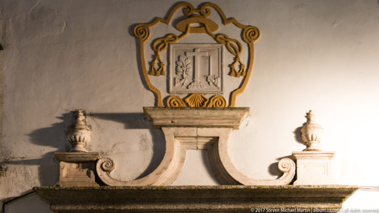 Illuminated stone heraldic cartouche above a curved basin, flanked by urn finials on a white wall.