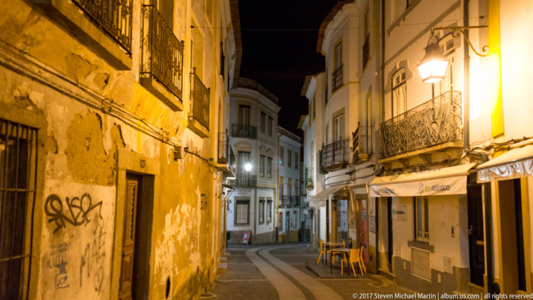 Narrow Évora street at night with whitewashed buildings, iron balconies, shopfronts, and patterned paving.