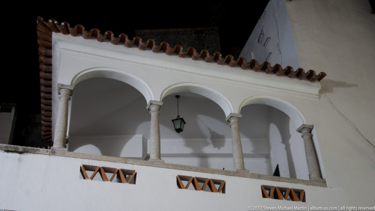 Whitewashed upper loggia with three rounded arches, slender stone columns, hanging lantern, and red-tile roof at night.
