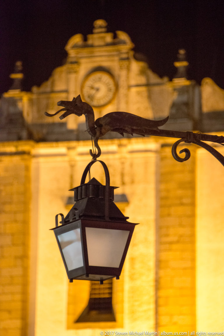 Street lantern suspended from a dragon-shaped metal bracket against a softly focused illuminated historic façade at night.