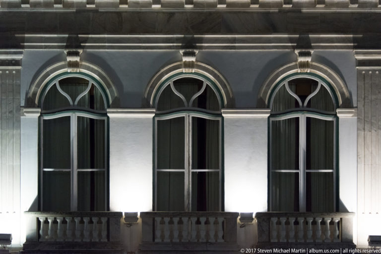 Three tall illuminated arched windows with stone surrounds and short balustrades on Évora's Banco de Portugal building.