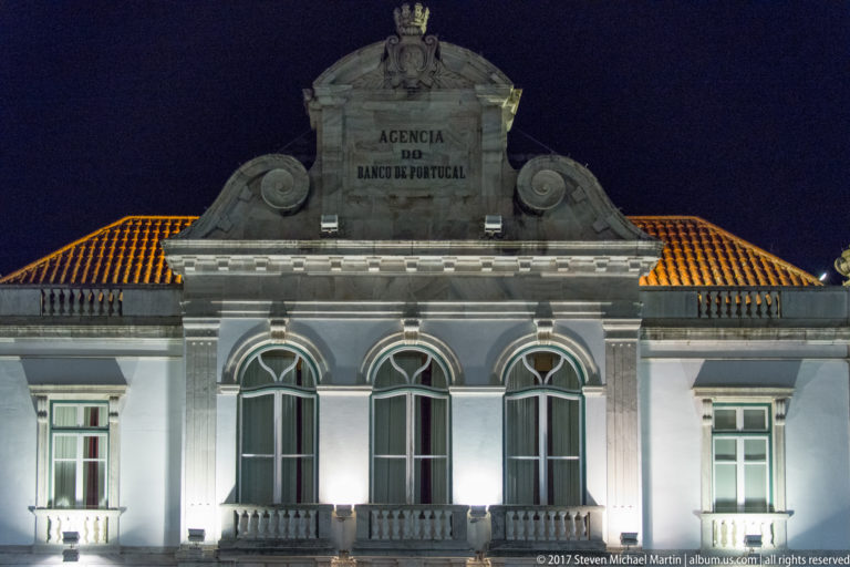 Night view of the Évora Banco de Portugal branch with three tall arched windows, balconies, and inscribed pediment.
