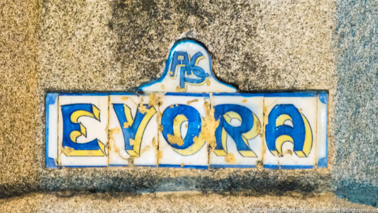 Small weathered ceramic plaque spelling Évora in blue-and-yellow letters on a rough stone wall.