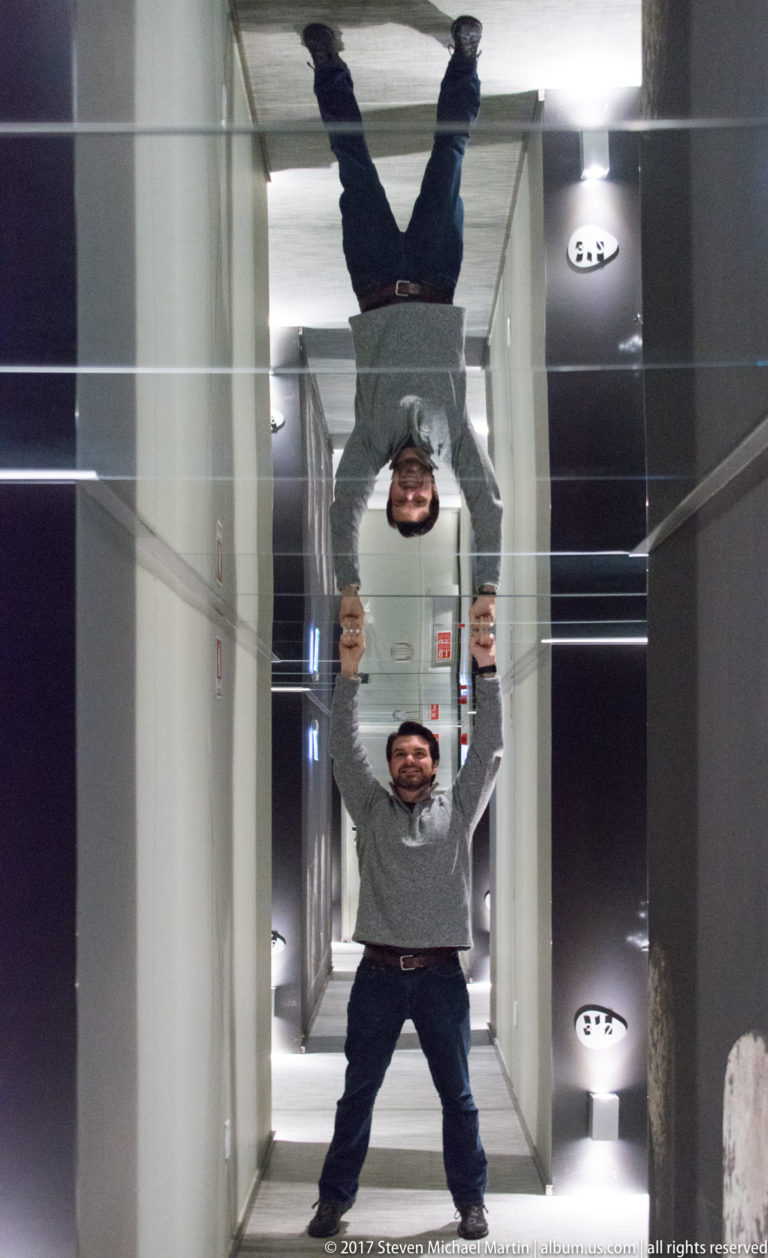 Visitor standing in a mirrored corridor with an inverted full-body reflection directly above him.
