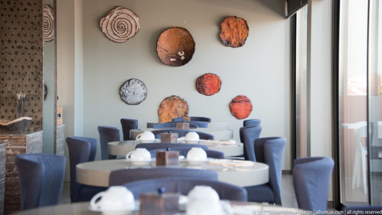 Modern dining room with round white tables, blue chairs, colorful ceramic wall pieces, and tall windows.