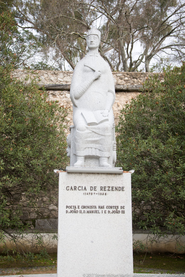 Stone statue of Garcia de Resende seated above an inscribed pedestal in a garden setting.