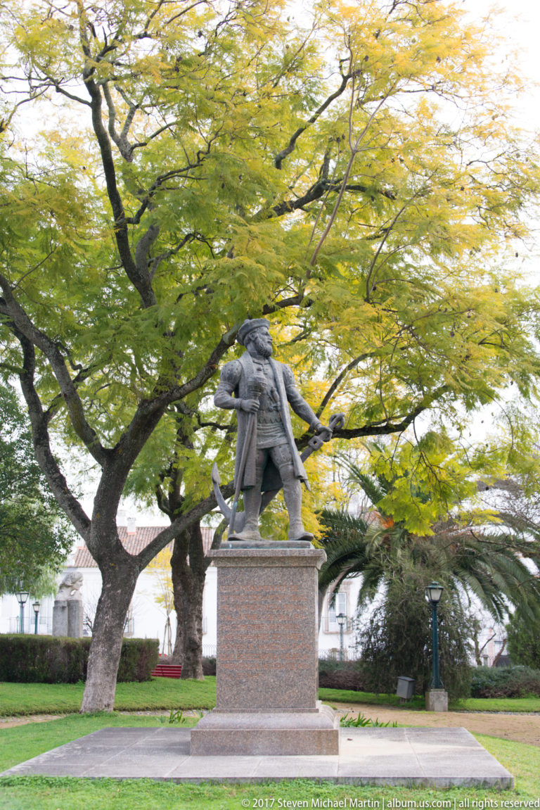 Bronze standing male figure on a tall stone pedestal beneath mature trees in an Évora garden.