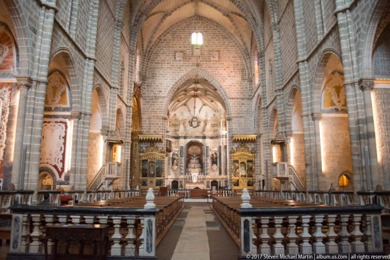 Centered church nave view toward a distant sanctuary, with rib vaults, stone piers, side altars, and foreground balustrade in the Igreja de São Francisco complex in Évora.