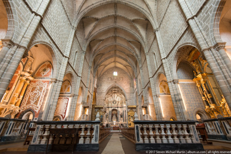Long Gothic church nave with pointed rib vaults, clustered stone piers, pews, and richly decorated side chapels in the Igreja de São Francisco complex in Évora.
