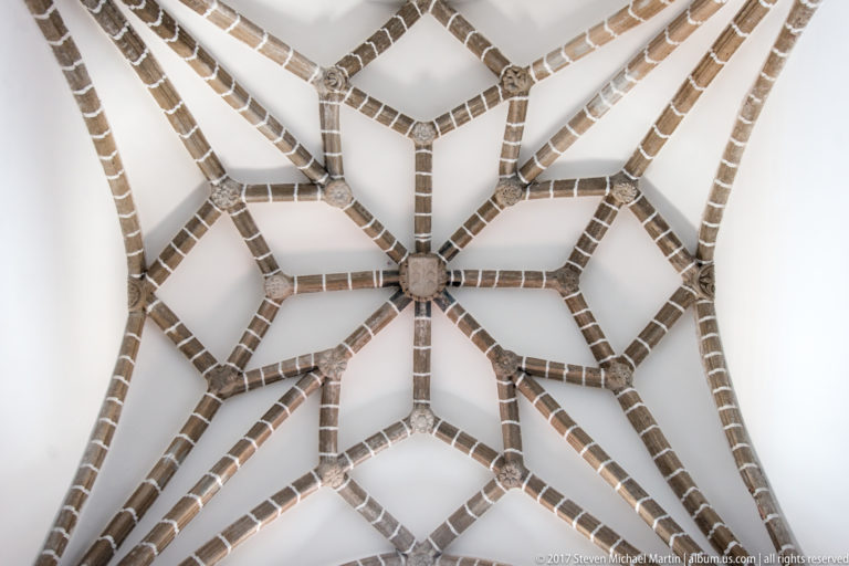 White ceiling crossed by dark stone ribs forming an elaborate star-shaped Gothic vault with a central boss.