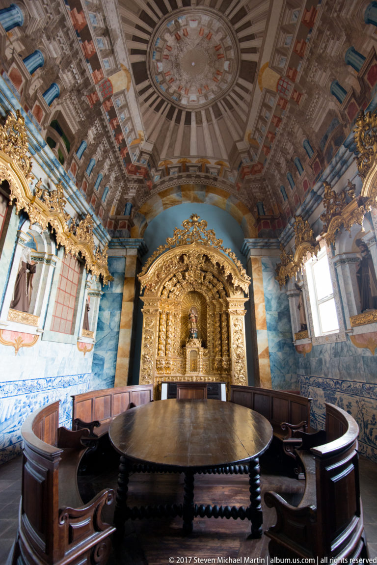 Decorated circular room with round table, wooden seating, blue-and-white tiles, painted ceiling, and gilded altar in the Igreja de São Francisco complex in Évora.