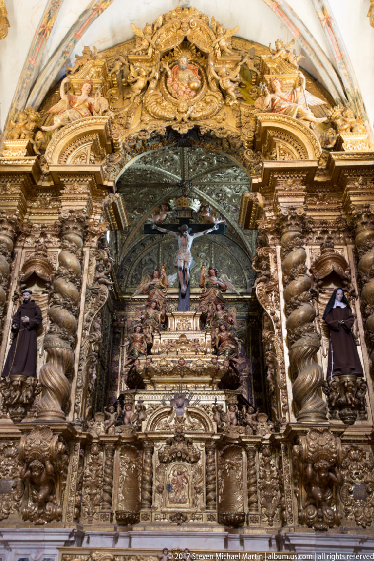 Dark Baroque retable with twisting columns, hooded Franciscan figures, gilded ornament, and a central devotional scene in the Igreja de São Francisco complex in Évora.