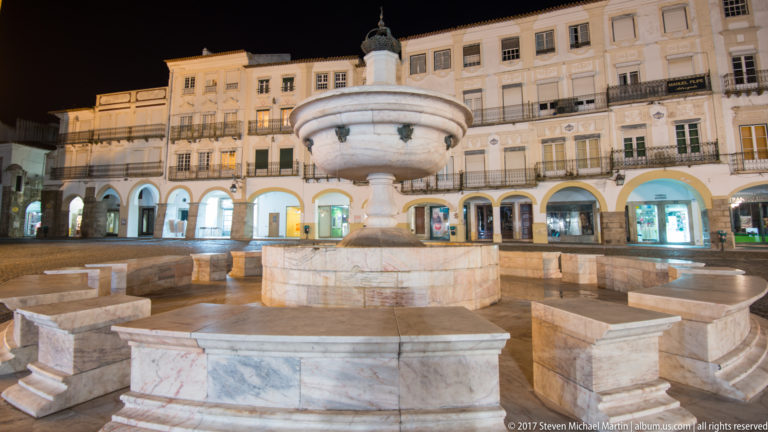 Marble fountain with octagonal basin in Praça do Giraldo at night, surrounded by arcaded historic façades.