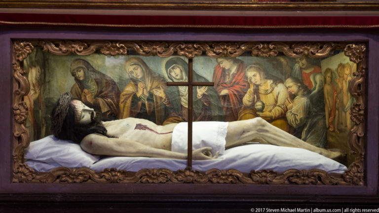 Reclining wounded figure of the dead Christ beneath glass, with painted mourners and a cross in the background.