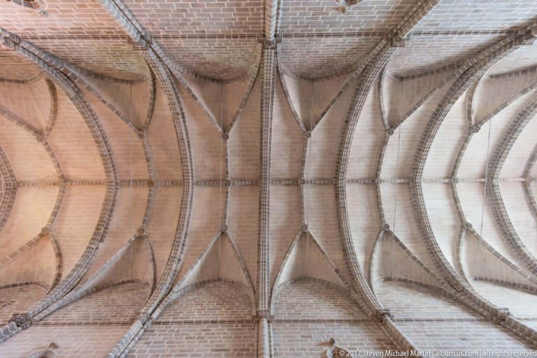 Upward view of repeated Gothic rib vaults in warm brick-colored masonry with pointed arches and intersecting ribs.