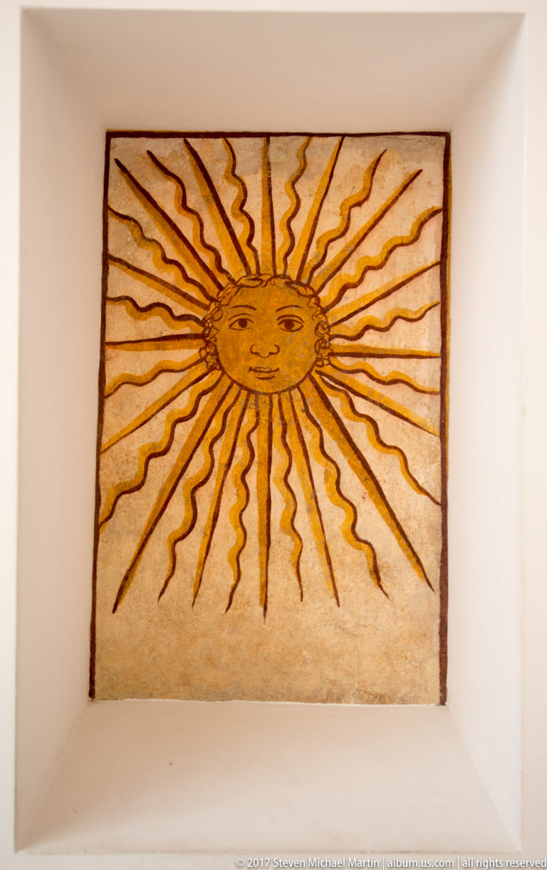 Rectangular ceiling panel painted with a golden human-faced sun and alternating straight and wavy rays.