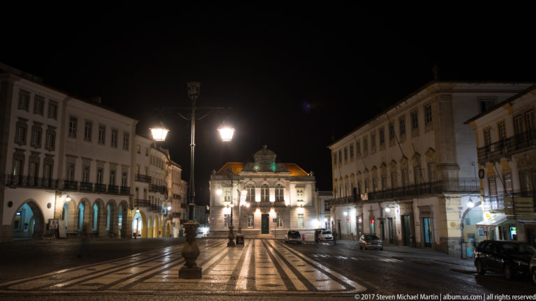 Striped paving leading across an Évora square toward the illuminated Banco de Portugal building, framed by historic façades and street lamps.