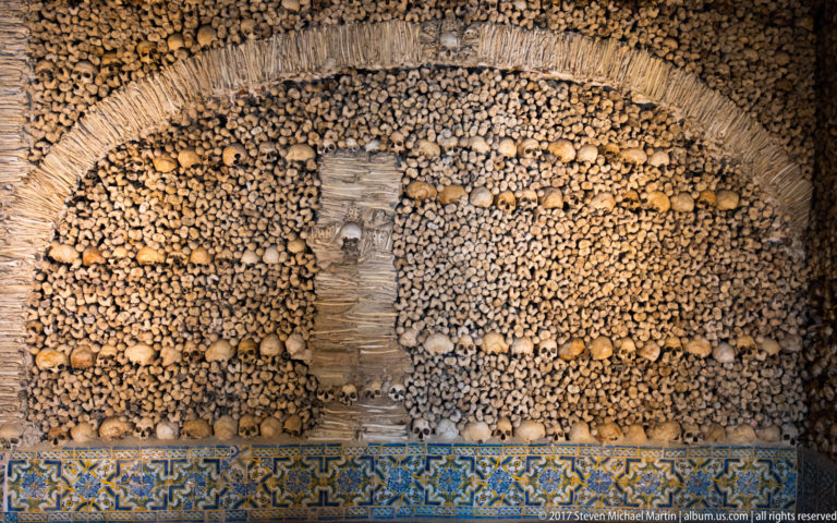 Arched ossuary panel of skulls and long bones above a blue-and-yellow tile border in Évora's Chapel of Bones.
