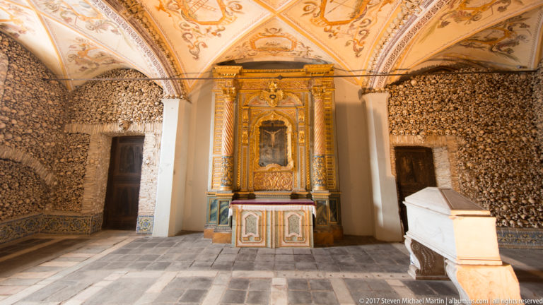 Gilded altar centered between bone-covered walls and beneath painted vaults inside Évora's Chapel of Bones.